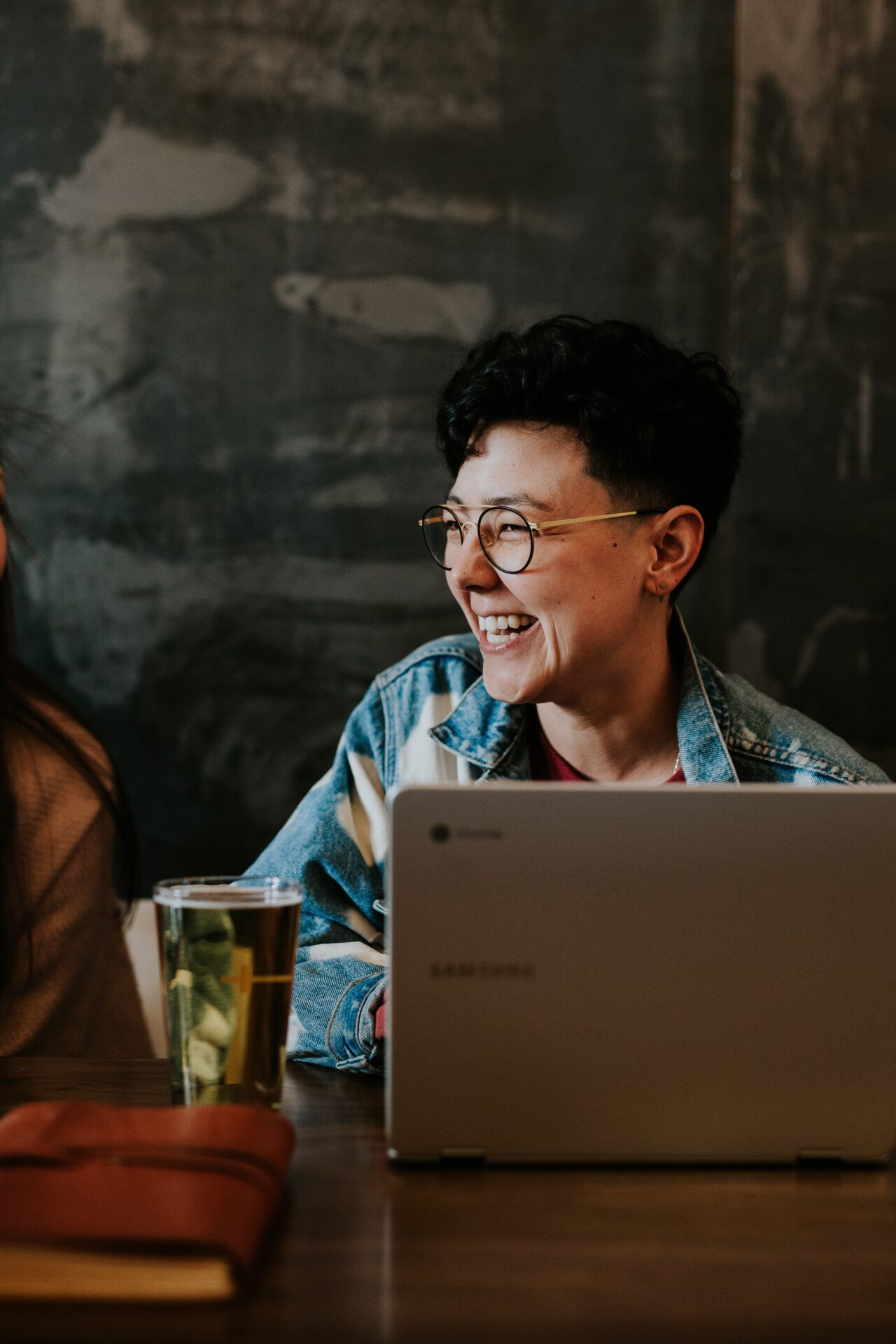 Person with glasses smiling with a drink and a laptop (Photo by Brooke Cagle on Unsplash)
