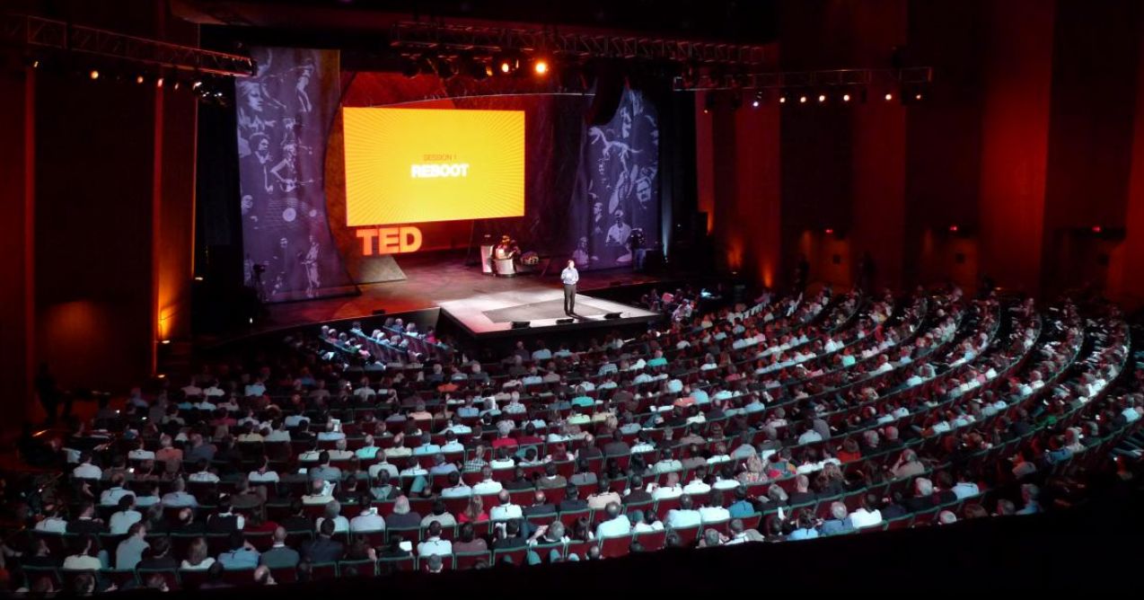 A wide shot of someone giving a TED talk to an audience