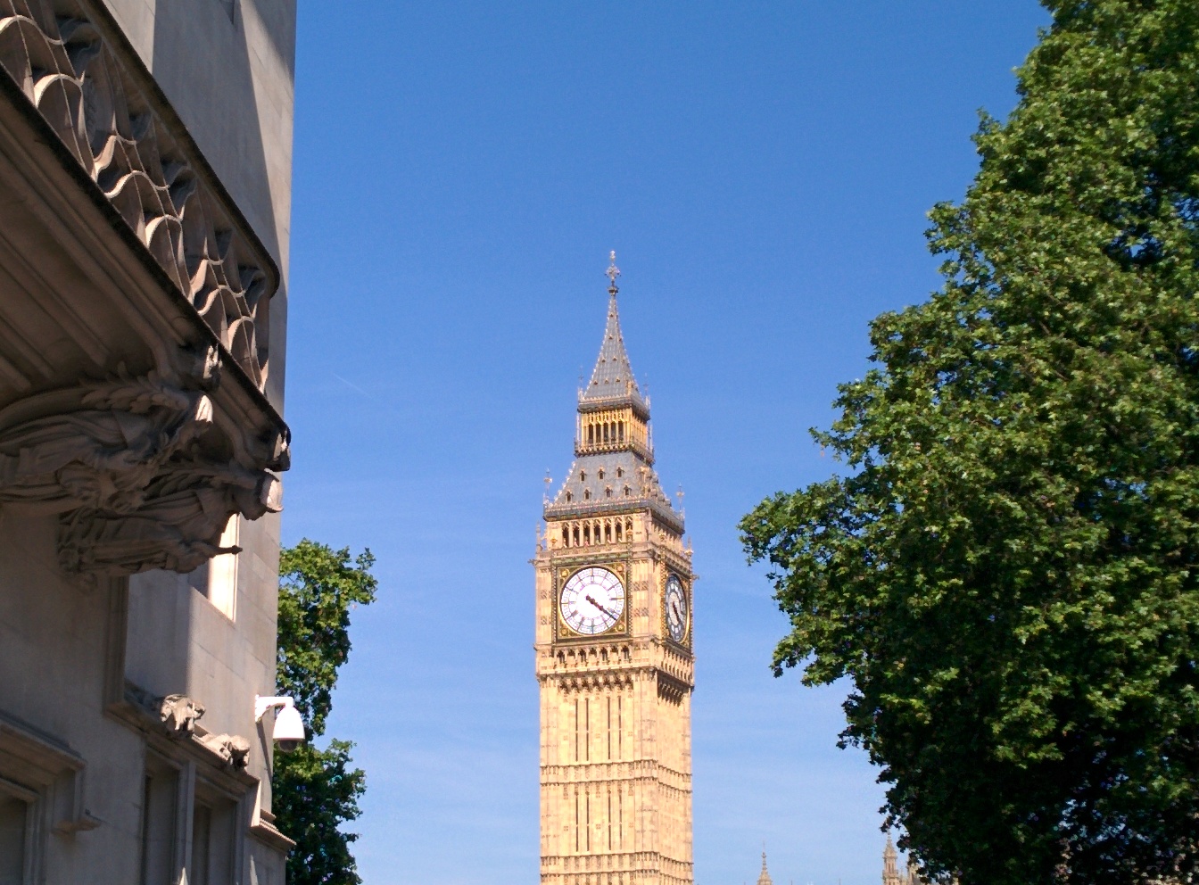 The Elizabeth Tower, housing the bell known as Big Ben, stands between a grey stone building and trees against a blue sky (Photo by Jock Busuttil / Product People Limited)