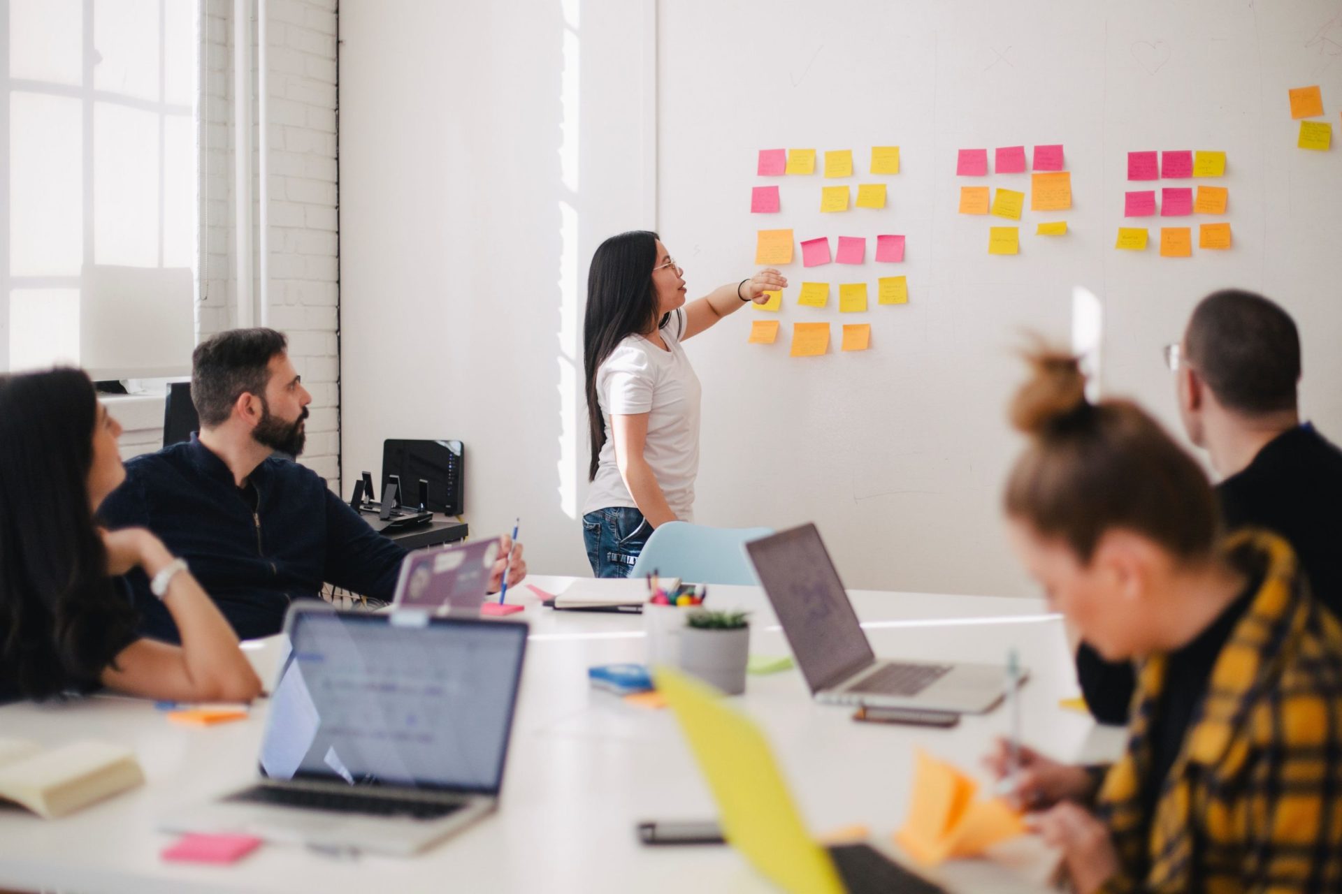 Woman in a tech startup presenting to her seated colleagues in front of a wall of colourful sticky notes. (Photo by You X Ventures on Unsplash)
