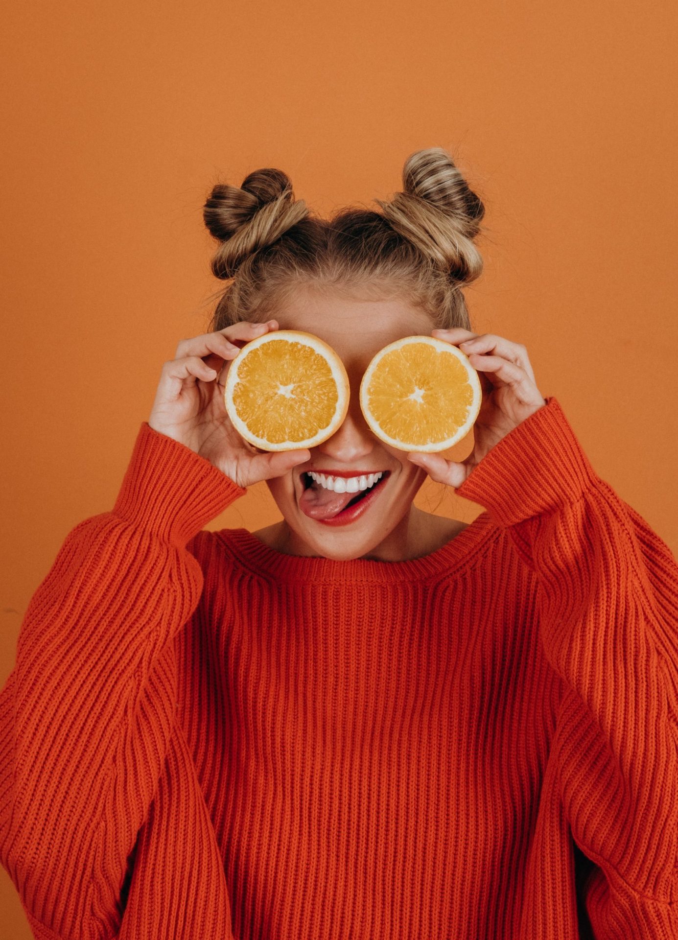 Woman in red knit sweater with bunches in her hair holds orange halves over her eyes while sticking out her tongue. Photo by Noah Buscher on Unsplash.