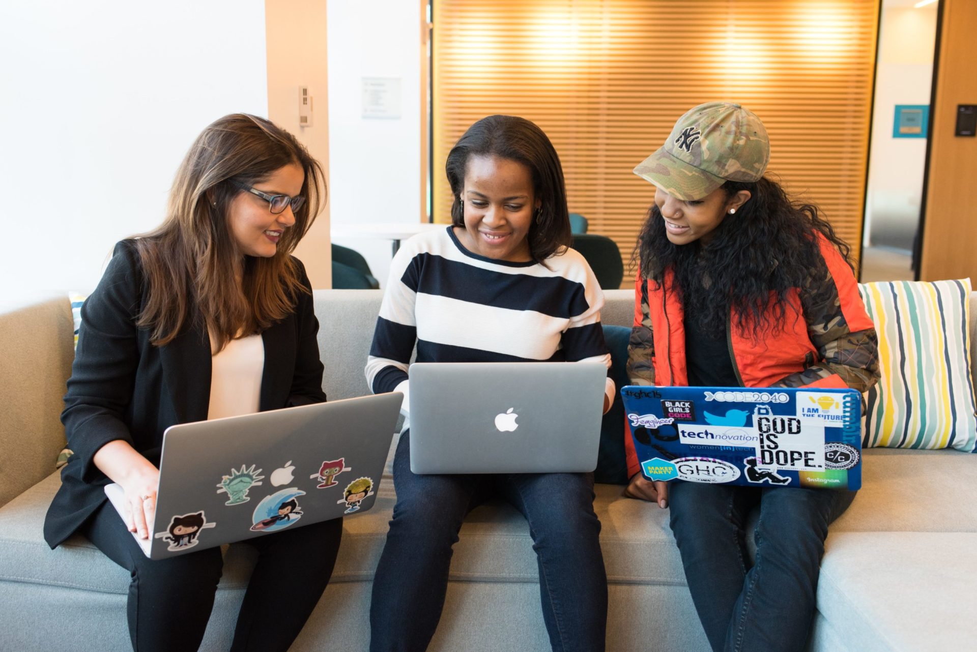 Three women sitting on a sofa with their MacBooks, which have varying numbers of techie stickers on them (Photo by Christina @ wocintechchat.com on Unsplash)