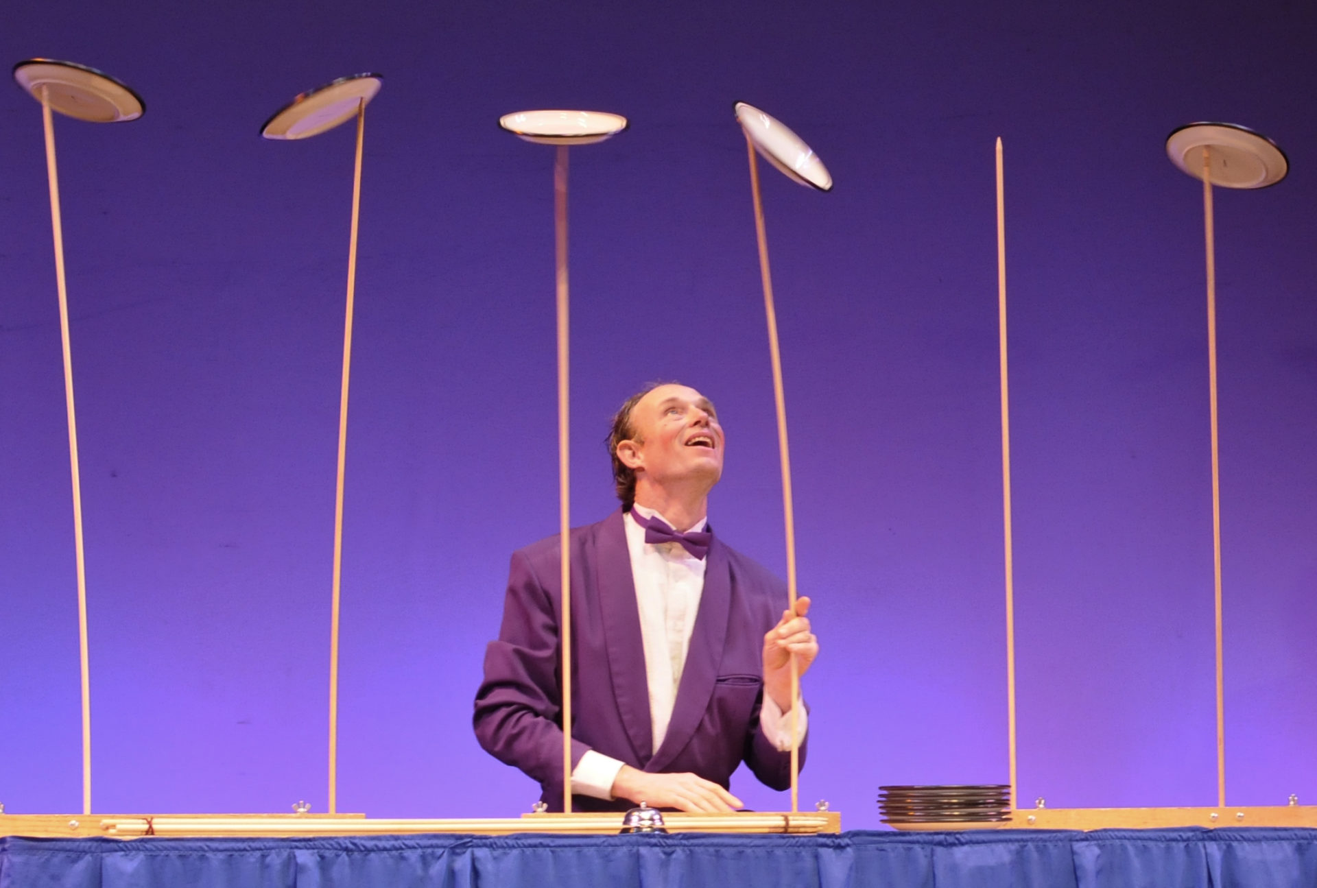 Man in purple jacket and matching bowtie spinning plates on tall sticks (Photo by Henrik Bothe / CC BY-SA)