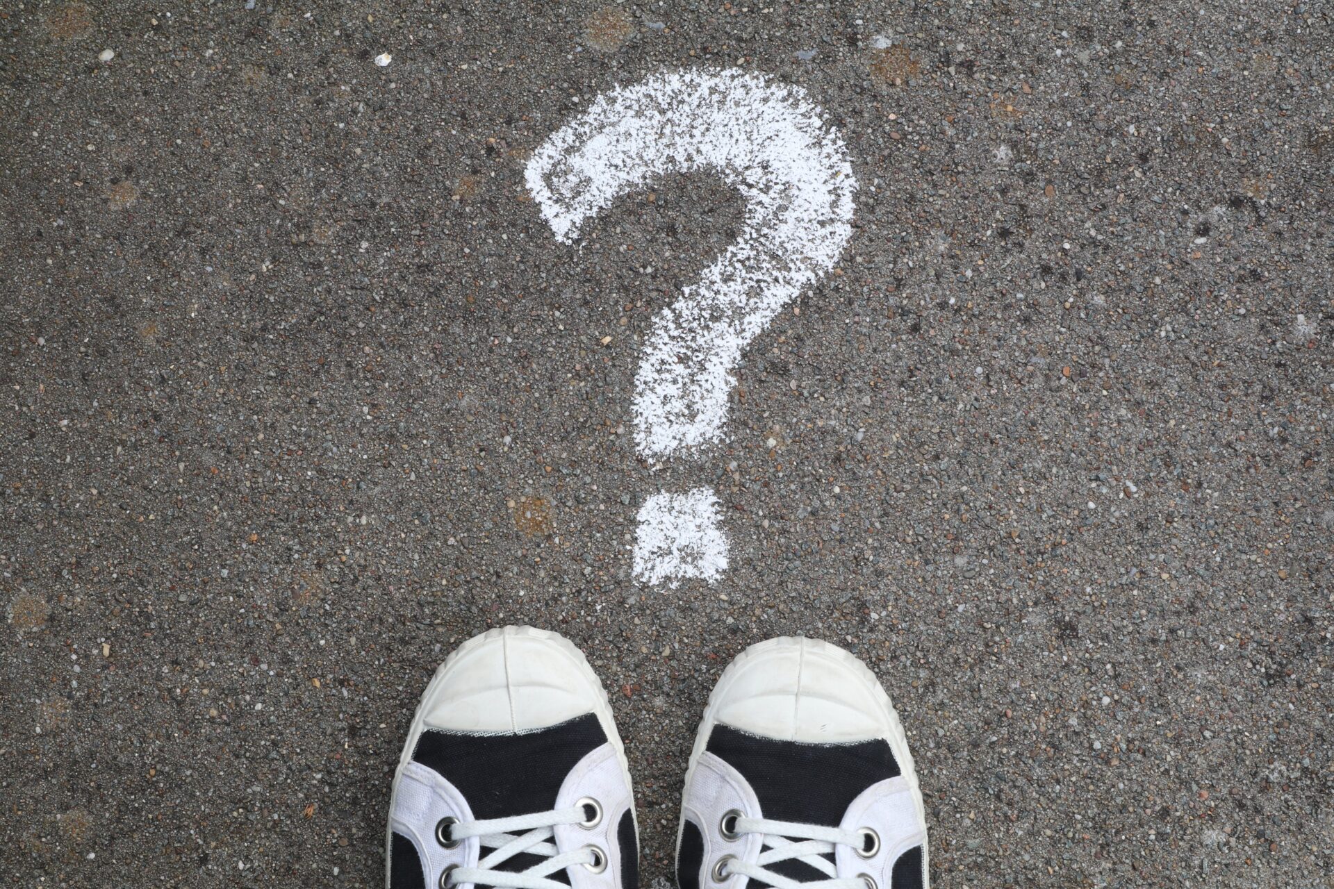 A question mark drawn in chalk on the concrete floor in front of a pair of feet in sports shoes. Photo by Ann H from Pexels